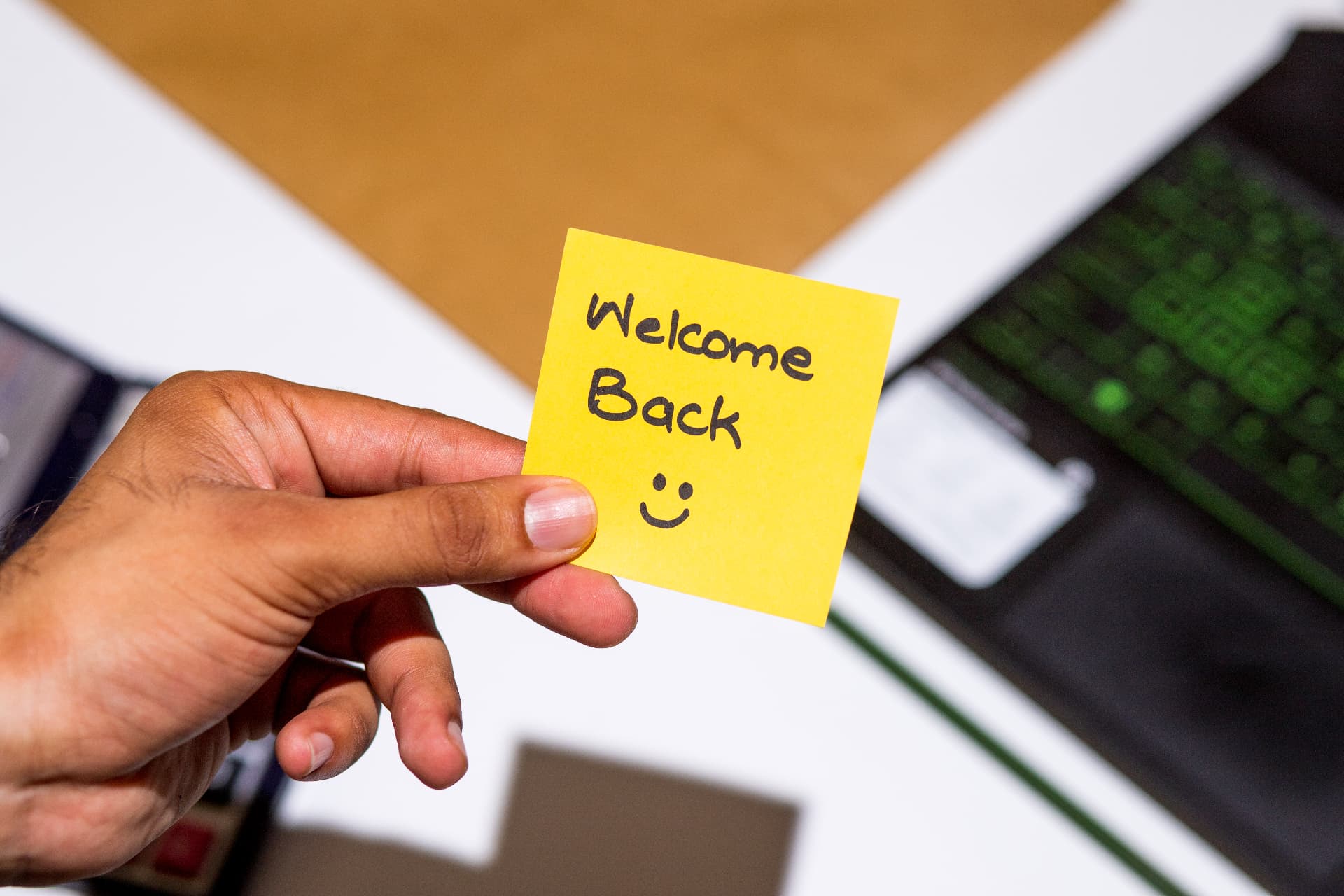 A hand holding a yellow sticky note that reads Welcome Back with a smiley face