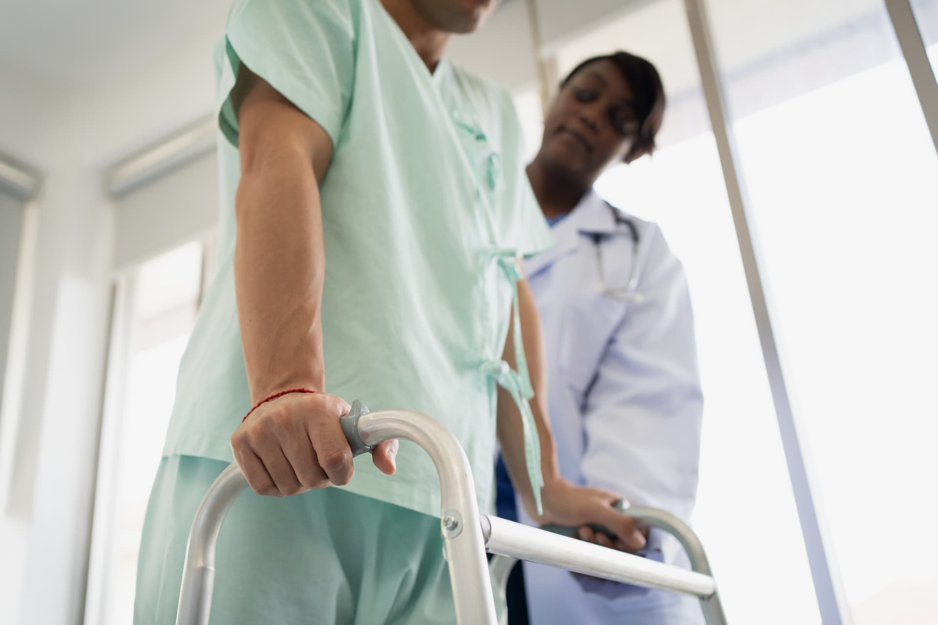 A patient in a hospital gown using a walker with a doctor watching in support